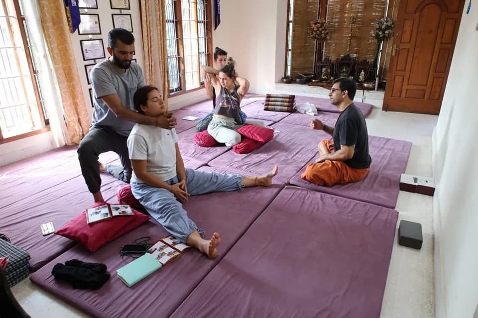 An instructor guiding a seated student's shoulder stretch while others practise on mats