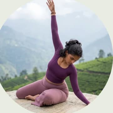 A woman in a side-bending pose on a stone ledge above tea hills